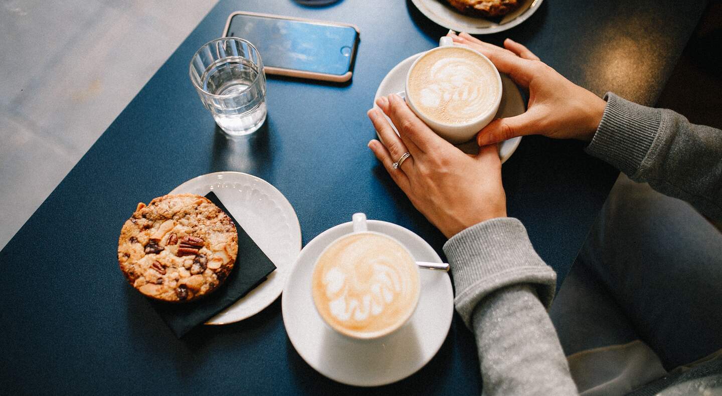 café faz mal? imagem mostra uma mesa com duas xícaras de café e parte das mãos de uma mulher segurando uma das xícaras.