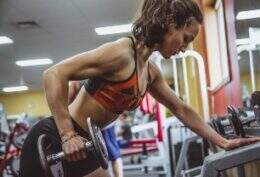 Imagem mostra mulher em ambiente de academia fazendo um treino de muculação. Ela tem os braços definidos e segura uma halter de anilha pequeno.