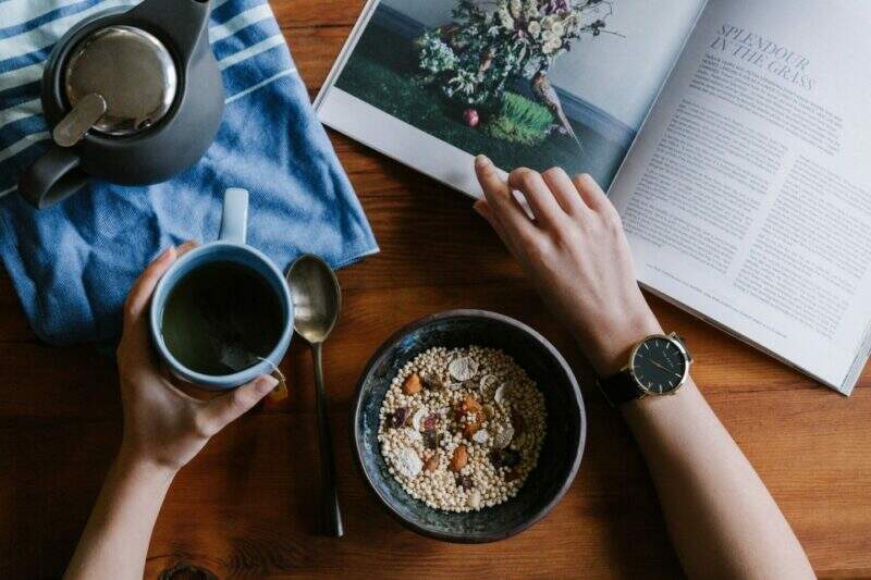 Pessoa com uma caneca de chá, um bowl de grãos para uma refeição saudável, folheando um livro sobre como criar hábitos