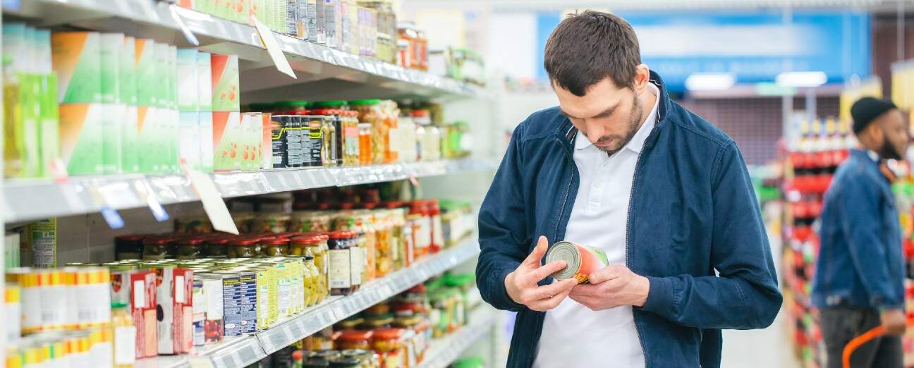 Em um corredor de supermercado, homem parado ao lado de uma prateleira segura nas mãos uma lata e lê atentamente o rótulo