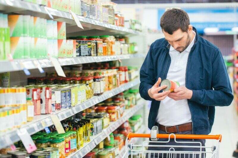 Em um corredor de supermercado, homem parado ao lado de uma prateleira segura nas mãos uma lata e lê atentamente o rótulo