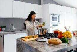 Imagem mostra uma mulher sorrindo enquanto, em uma cozinha grande e bem iluminada, prepara alimentos naturais e muito coloridos