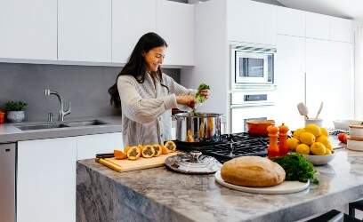 Imagem mostra uma mulher sorrindo enquanto, em uma cozinha grande e bem iluminada, prepara alimentos naturais e muito coloridos