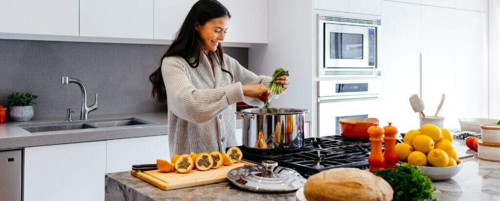 Imagem mostra uma mulher sorrindo enquanto, em uma cozinha grande e bem iluminada, prepara alimentos naturais e muito coloridos