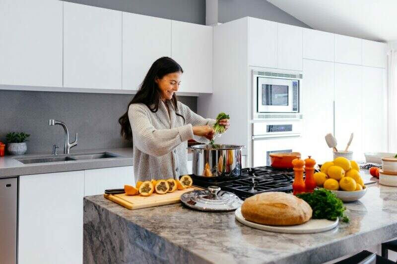 Imagem mostra uma mulher sorrindo enquanto, em uma cozinha grande e bem iluminada, prepara alimentos naturais e muito coloridos
