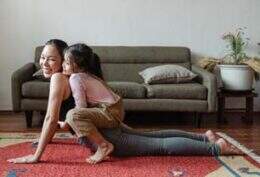 Imagem mostra mulher tentando praticar exercícios físicos no tapete de uma sala de estar, enquanto uma criança brinca sentada sobre suas costas, com as pernas ao redor da cintura da mãe