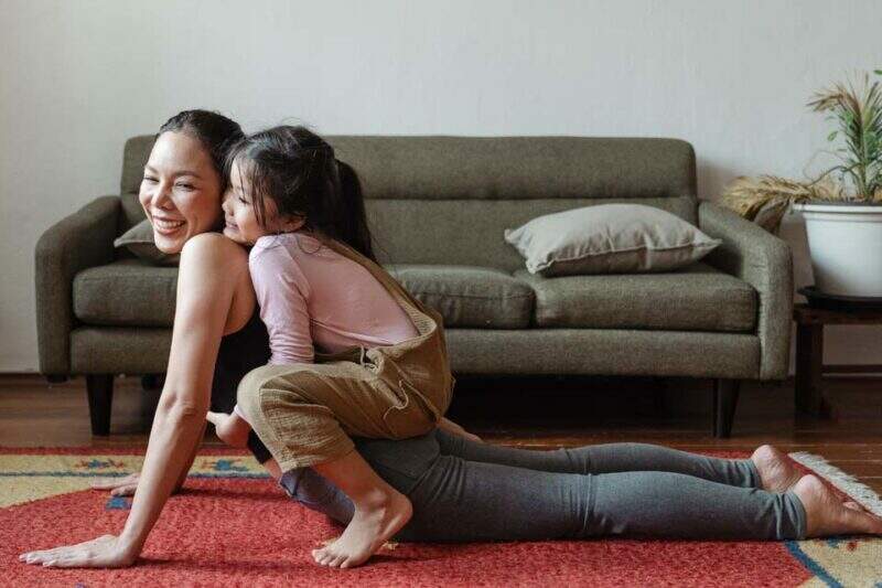 Imagem mostra mulher tentando praticar exercícios físicos no tapete de uma sala de estar, enquanto uma criança brinca sentada sobre suas costas, com as pernas ao redor da cintura da mãe