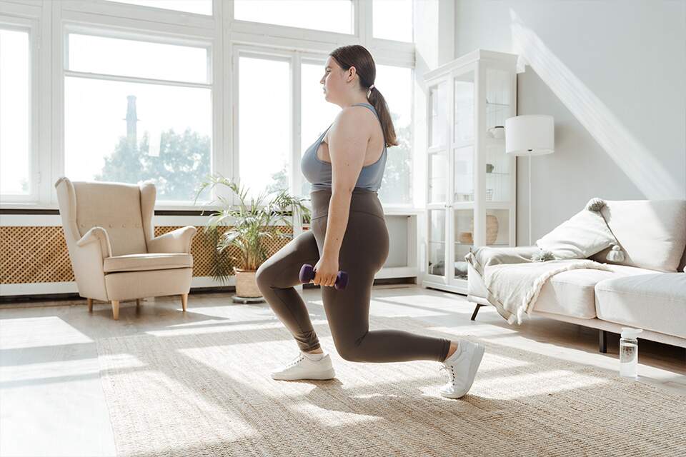 Imagem mostra mulher praticando afundo na sala de uma casa