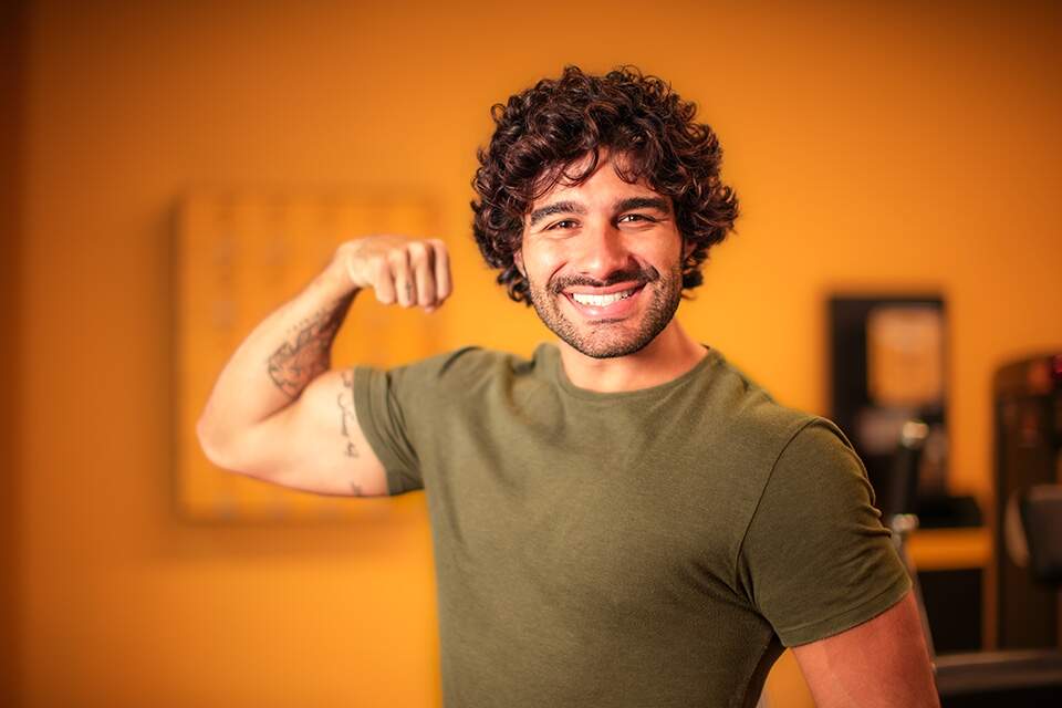 Imagem mostra homem sorrindo e mostrando o bíceps em ambiente de academia