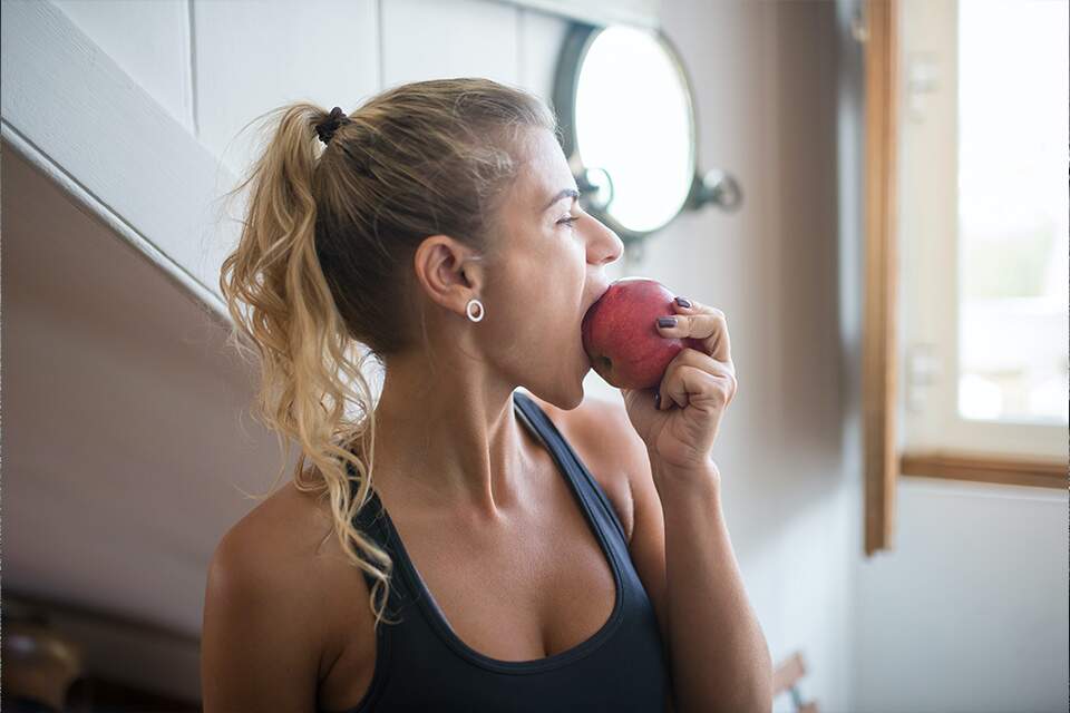 Na imagem, mulher em roupas de treino come uma maçã