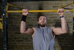 Imagem mostra homem praticando Pull-UP assistido em ambiente de academia