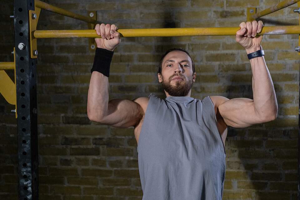 Imagem mostra homem praticando Pull-UP assistido em ambiente de academia