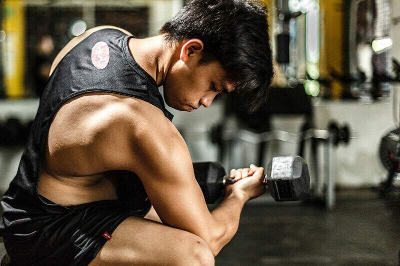 Imagem mostra homem em em ambiente de academia, com o braço flexionado, treinando com um haltere