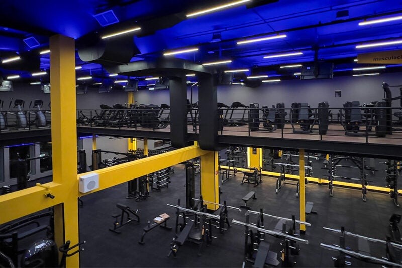 Vista interna da academia Smart Fit com área de musculação e cardio em dois andares, equipada e iluminada em tons de azul e amarelo.