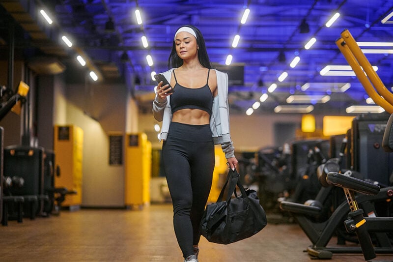 Mulher com roupa de treino caminhando pela academia Smart Fit com bolsa esportiva e celular na mão, ambiente moderno e iluminado em tons de azul e amarelo