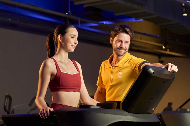 Pessoa recebendo orientação de instrutor da Smart Fit enquanto utiliza a esteira na academia, em ambiente moderno e iluminado.