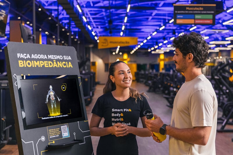 Pessoa recebendo orientação sobre bioimpedância na academia Smart Fit ao lado da máquina de avaliação corporal, em ambiente moderno e iluminado.