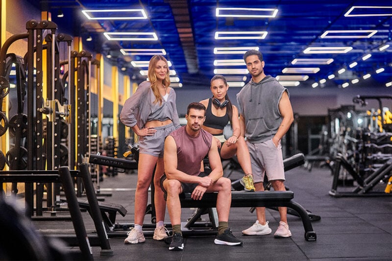 Grupo de pessoas posando na área de peso livre da academia Smart Fit, com equipamentos ao fundo em ambiente iluminado.