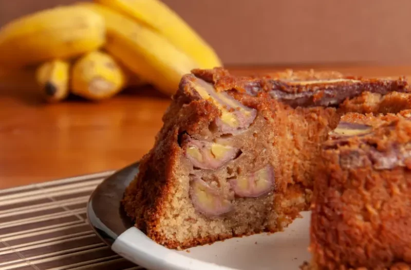 imagem de um prato com um bolo de banana cortado e um cacho de bananas ao fundo, em cima de uma mesa de madeira.