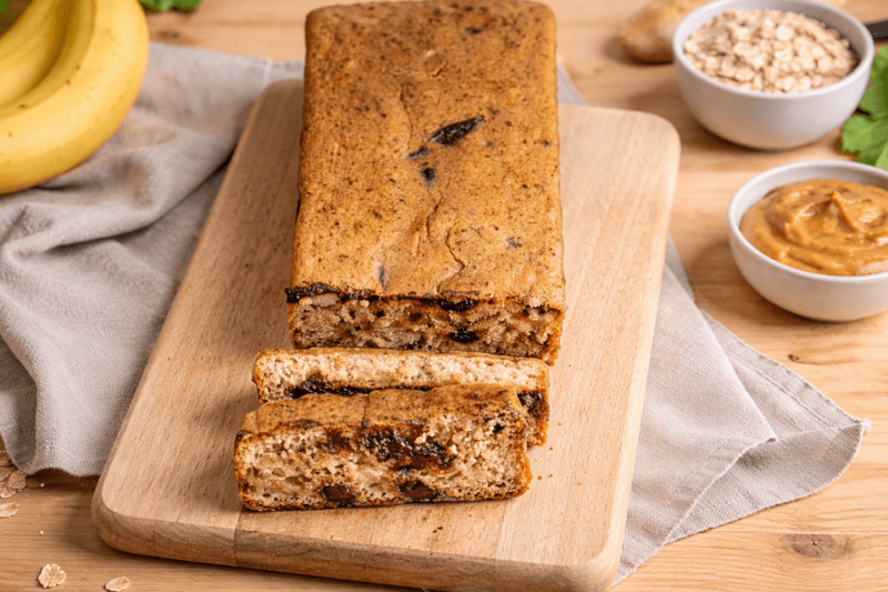 imagem de um prato com um bolo de banana fit cortado e um cacho de bananas ao fundo, em cima de uma mesa de madeira.